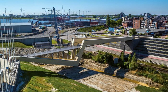 Die neue Fußgänger- und Radweg-Brücke verbindet die Stadt Delfzijl mit dem Wattenmeer. Ihre markante Z-Form nimmt die Gegebenheiten vor Ort auf: Mit dem oberen Schenkel überbrückt sie als geschlossener Trog Straße und Bahntrasse, mit dem unteren bildet sie die Topographie des Deichs nach und führt auf dessen Krone.