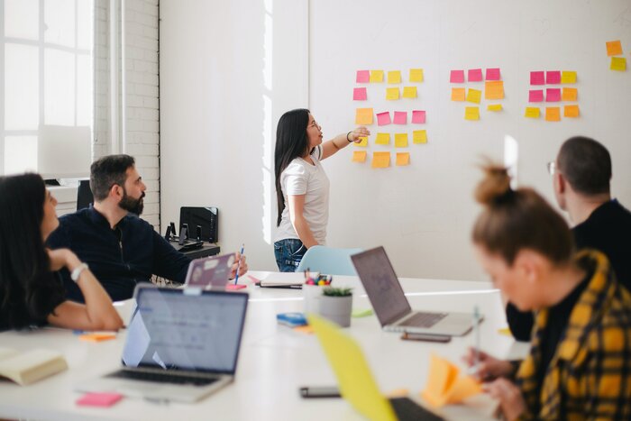 Team im Büro vor Laptop an einem großen Schreibtisch, eine Frau zeigt auf Post its am Boad