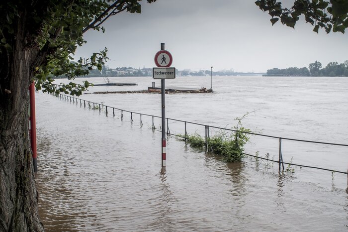 Uferpromenade eines Flusses in Deutschland steht unter Wasser, ein Schild "Hochwasser" im Zentrum des Bildes