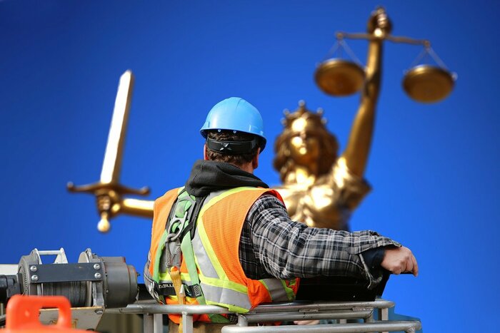 Ein Bauarbeiter mit Helm schaut auf eine goldene Justizia-Figur vor blauem Himmel