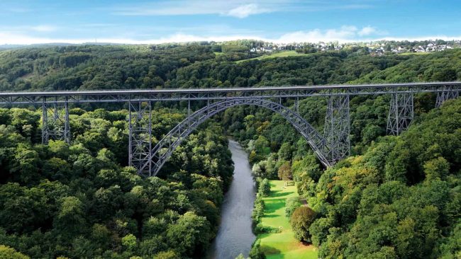 Auf gut 100 Metern Höhe spannt sich der parabelförmige Bogen über das tief eingeschnittene Tal der Wupper. Ansicht der Müngstener Brücke mit Brückenpark. Drohnenaufnahme September 2025 © Hello Studio W