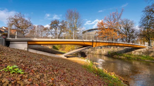 Bild 1: Die Lindachbrücke in Schwäbisch Hall überspannt den Kocher als elegantes Bauwerk. Die neue Fuß- und Radwegbrücke – eine Holz-Beton-Verbund-Konstruktion – bietet nun eine direkte Anbindung für Fußgänger und Radfahrer zwischen Bahnhof und der Unterwöhrd-Insel, auf der auch das Globe-Theater steht (im Hintergrund zu sehen), das damit ebenfalls den erforderlichen Zugang erhält. © Schaffitzel Holzindustrie – Jürgen Weller Fotografie