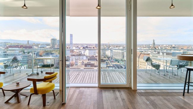 In den Attikageschossen unter den Flachdächern bieten Rundumverglasungen mit raumhohen Türen einen Blick auf die Stadt; im Neubau finden Angestellte hier oben informelle Aufenthaltsbereiche. © Zejlko Gataric, Zürich / Schüco International KG