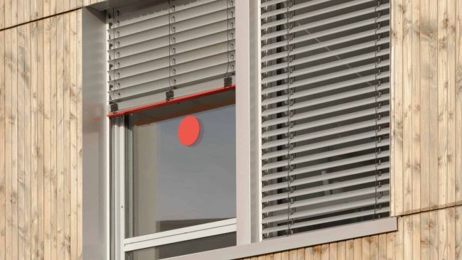 SecuKit ist eine Sicherheitsausstattung von Warema, mit der die Raffstoren manuell nach oben geschoben werden können. Die Endschiene arretiert sich auf vordefinierter Höhe seitlich in den Führungsschienen. Die so ausgestatteten Notausgänge sind mit roten Aufklebern auf der Scheibe gekennzeichnet. Das und die in Verkehrsrot gehaltenen Endschienen der Raffstoren machen die Rettungswege gerade dann schnell und klar erkennbar, wenn jede Sekunde zählt. © WAREMA