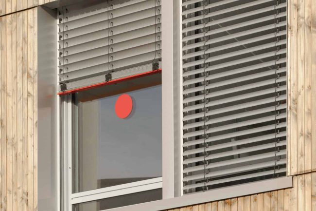 SecuKit ist eine Sicherheitsausstattung von Warema, mit der die Raffstoren manuell nach oben geschoben werden können. Die Endschiene arretiert sich auf vordefinierter Höhe seitlich in den Führungsschienen. Die so ausgestatteten Notausgänge sind mit roten Aufklebern auf der Scheibe gekennzeichnet. Das und die in Verkehrsrot gehaltenen Endschienen der Raffstoren machen die Rettungswege gerade dann schnell und klar erkennbar, wenn jede Sekunde zählt. © WAREMA